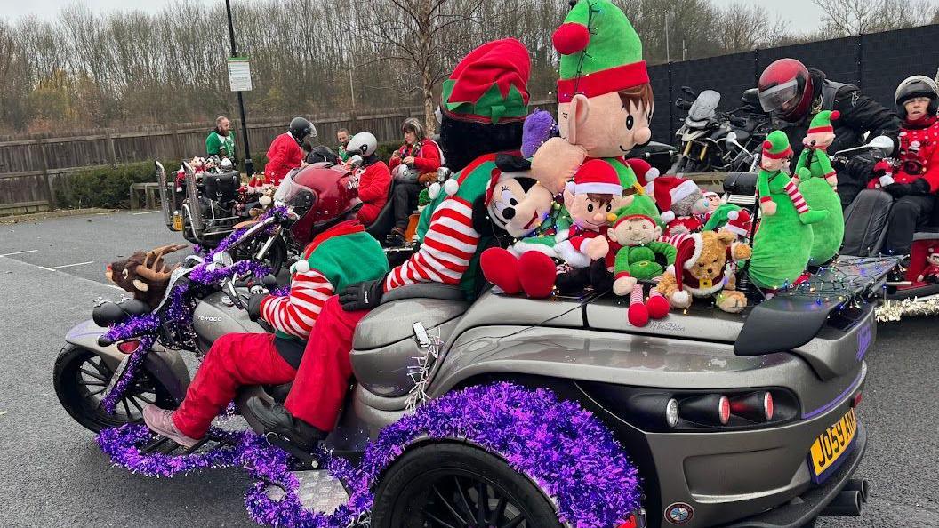 Motorcyclists sitting on a silver trike covered with giant soft toys and purple tinsel