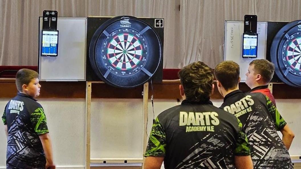 Four teenagers with Darts Academy North East shirts have their back to the camera and are throwing darts at a dart board. 