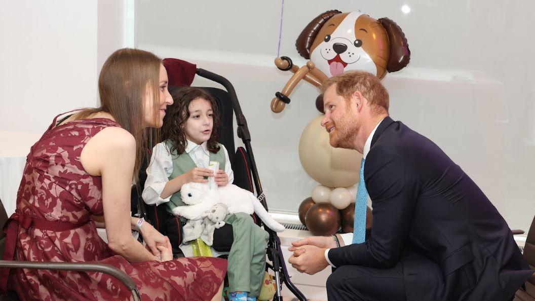 Prince Harry sat down facing and smiling at Noah's mum who is wearing a red dress and smiling back with Noah sat in the middle holding a white cat teddy