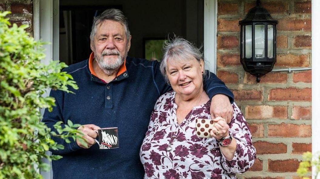 Lol and Sue Hepworth are standing outside their home and they are both holding a mug each. Lol is wearing a navy jumper and Sue is wearing a flowery shirt.