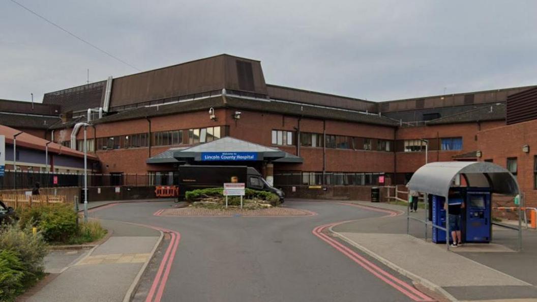 The main entrance to Lincoln County Hospital. A three-storey, red brick-built building. A large blue and white sign, in front of the building, states 'Lincoln County Hospital'