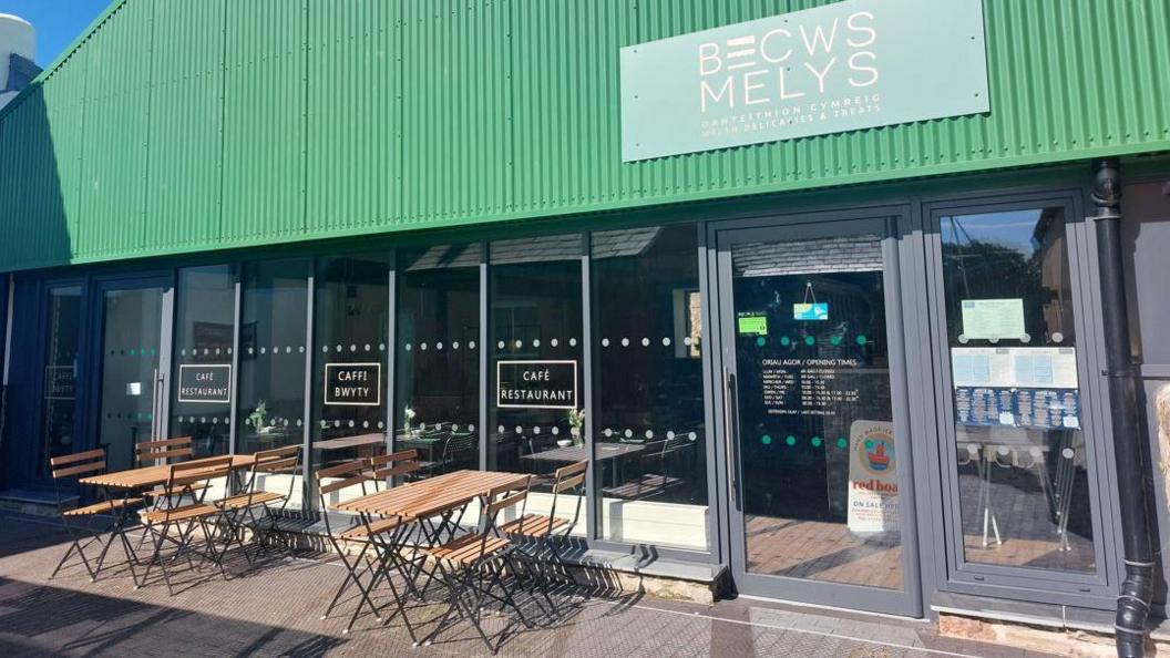 Becws Melys, in Caernarfon, is a restaurant with a green metal and glass frontage and tables and chairs in front of the building