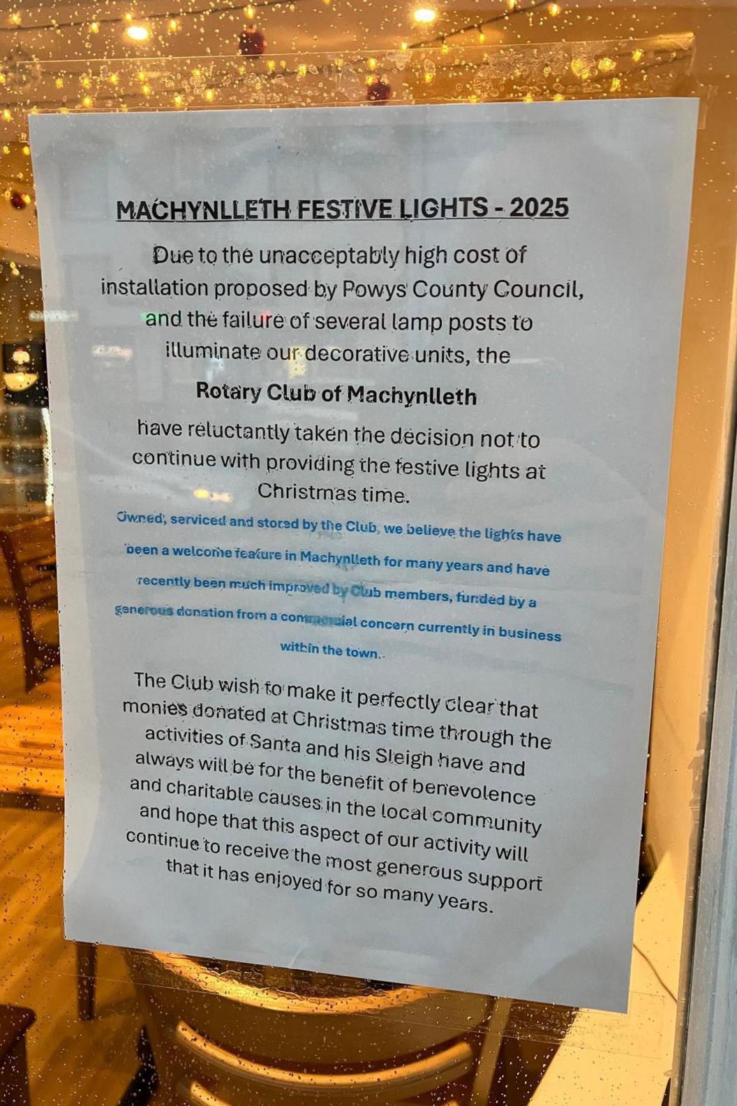 A piece of paper on a shop window. It says 'Machynlleth Festive Lights - 2025' in bold capitals. It continues 'Due to unacceptably high cost of installation proposed by Powys County Council, and the failure of several lamp posts to illuminate our decorative units, the Rotary Club of Machynlleth, have reluctantly taken the decision not to continue providing the festive lights at Christmas time.' It adds that the lights have been a feature feature funded by donations. It also states that money donated through the activities of Santa and his Sleigh have always been donated to local charities. 