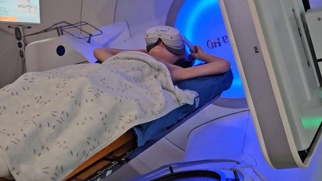 Coby is lying down in a radiotherapy machine. He is covered by a blanket and is wearing a VR headset.