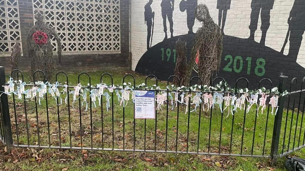 Railings with blue and pink ribbon and writing attached with a sign and barcode to scan in the middle in front of a remembrance sculpture of two people and a mural with soldiers on it in the background