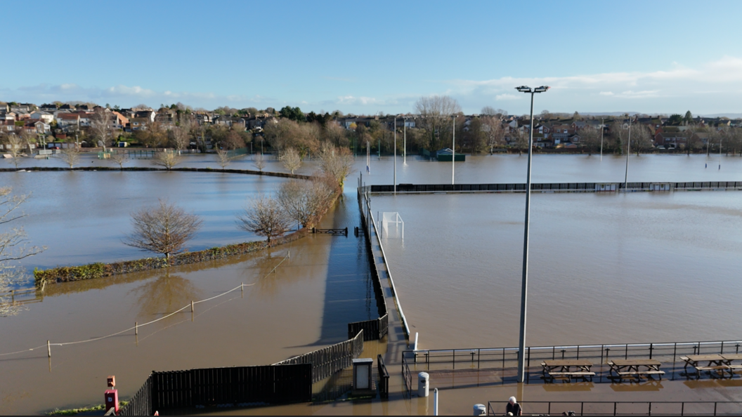 The football pitches pictured completely flooded, a long with fields surrounding it. A goal is submerged on the right-hand side of the photo and hedges and trees rise from the floodwaters.