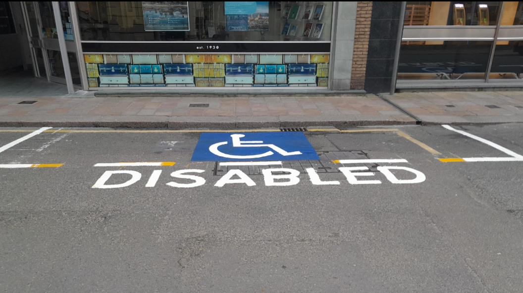 A parking space with fresh painting of a disabled blue symbol with DISABLED painted in white below on the road, with a stone paved path behind and buildings.