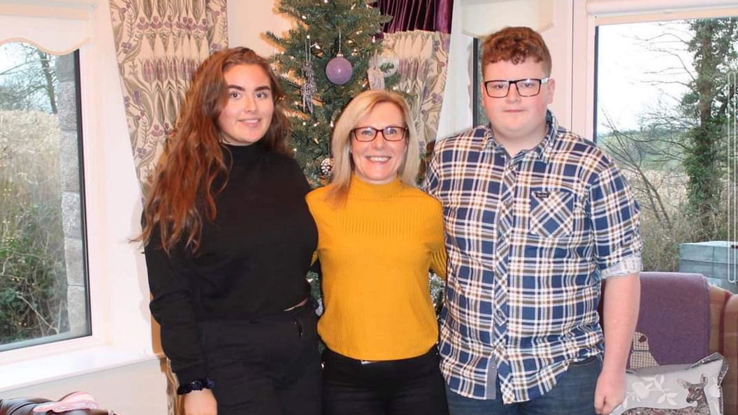 Jeni, Sandra and Daniel Larmour stood in front of a Christmas tree. Jeni is wearing black, Sandra is wearing a mustard top and glasses and Daniel is wearing a blue checked shirt with glasses.