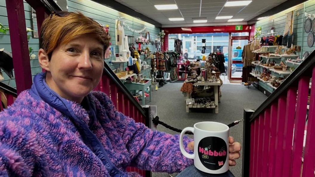 Dynes gyda gwallt byr yn gwisgo siwmper piws a pinc, yn eistedd ar risiau tu fewn i siop, yn dal paned yn ei llaw. Mae hi'n edrych at y camera ac yn gwenu. Mae gweddill y siop yn y cefndir. Mae'r grisiau yn binc.