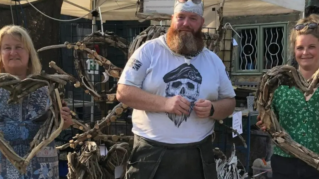 Dan Wakeman wearing a white t shirt. This has a skull on it wearing a military style cap. Dan is wearing a white bandana with black dot detailing. Two other people are next to him holding driftwood heart sculptures.