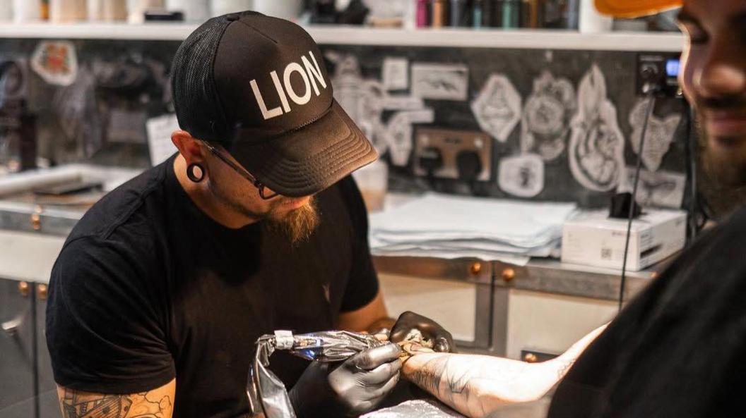 A man is wearing a black t-shirt and a black cap. He is holding a tattoo gun as he tattooes a man's arm.