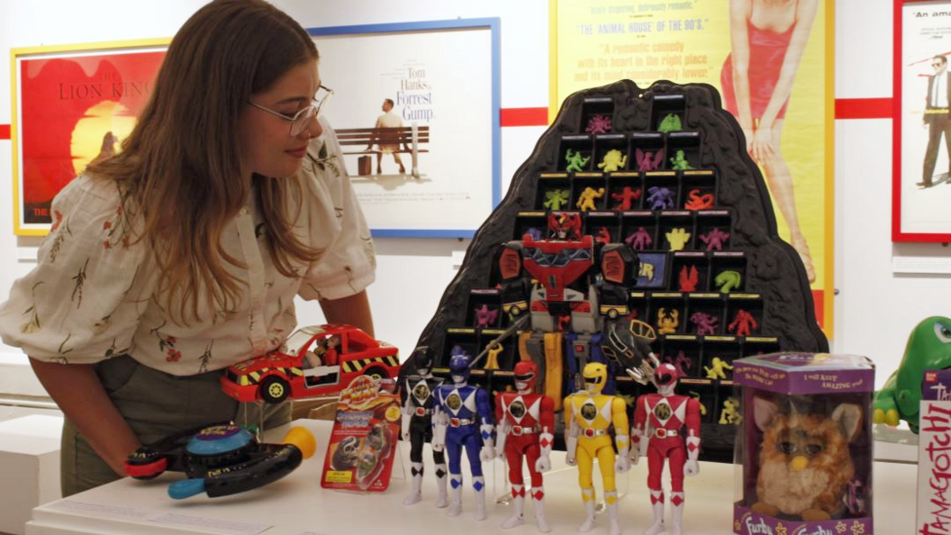 A woman stands and looks at a selection of the toys on display, including Power Rangers, Furby and Tamagotchi. Music posters can be seen in the background.