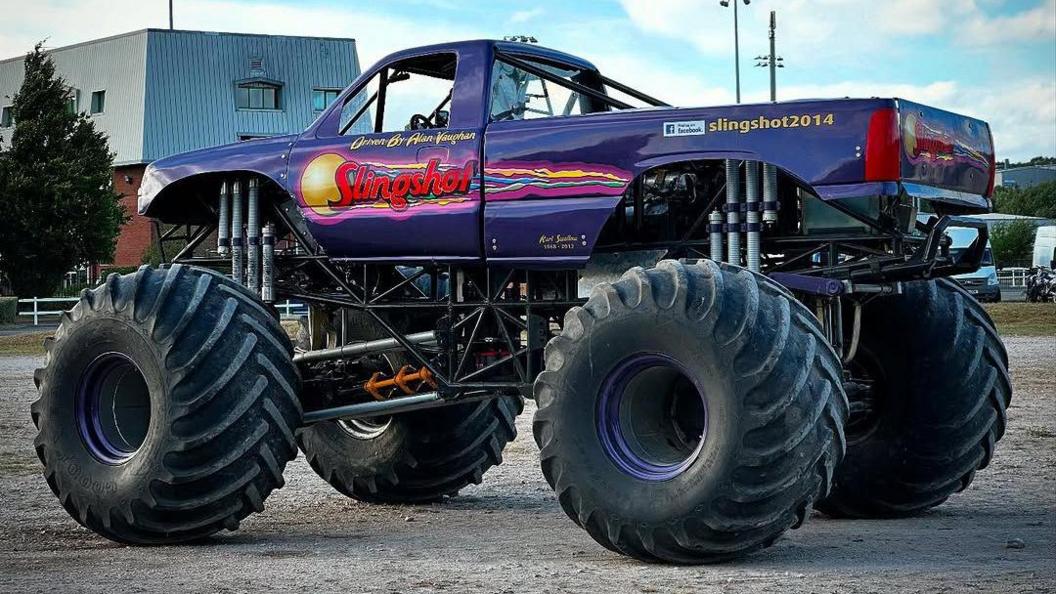 A purple monster truck is driving on a field. It has the word 'Slingshot' on the side of the truck.