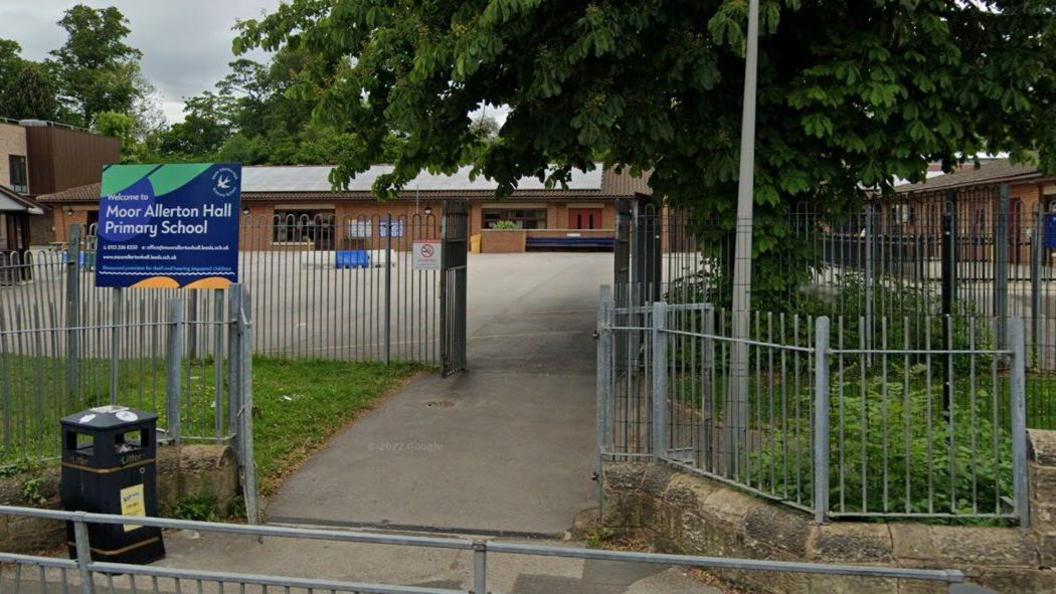 The metal fenced entrance to Moor Allerton Hall Primary School with a blue sign on the left which features the school's name