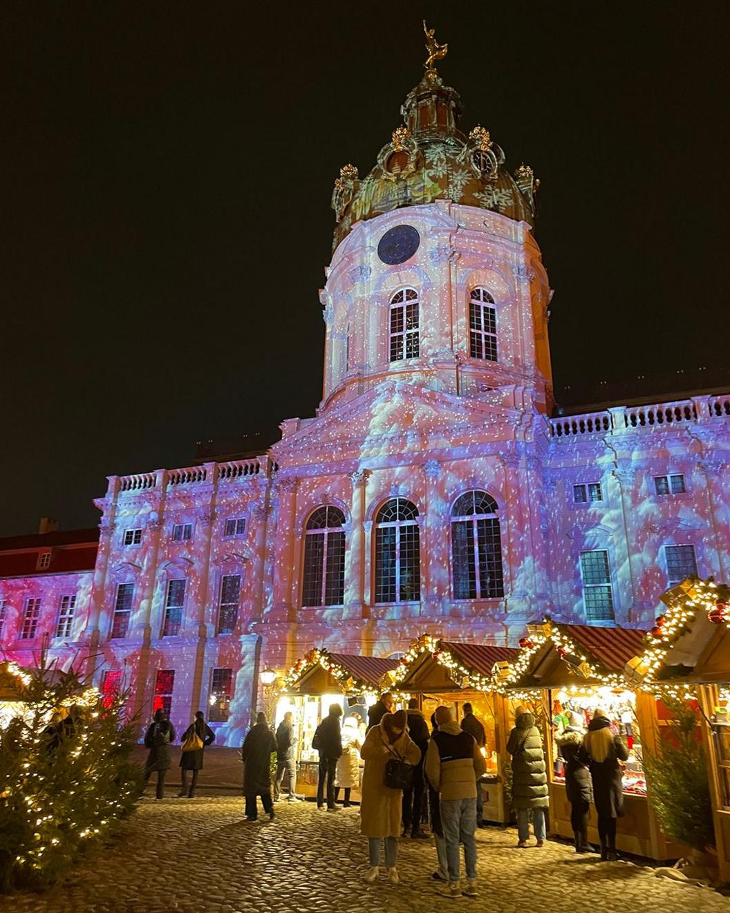A pink projection onto a historical palace building in Berlin, Germany, with wooden huts lined with fairy lights and Christmas trees in the foreground in front of the palace, as people mingle at Christmas market stalls.