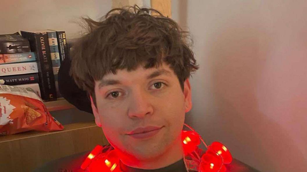 Luis Piovesana wearing a dark grey jumper and sitting down in a corner, leaning up against a wooden bookcase. He has a string of red, love-heart shaped fairy lights around his neck. He has a dark brown, shaggy haircut and brown eyes, and is softly smiling at the camera.