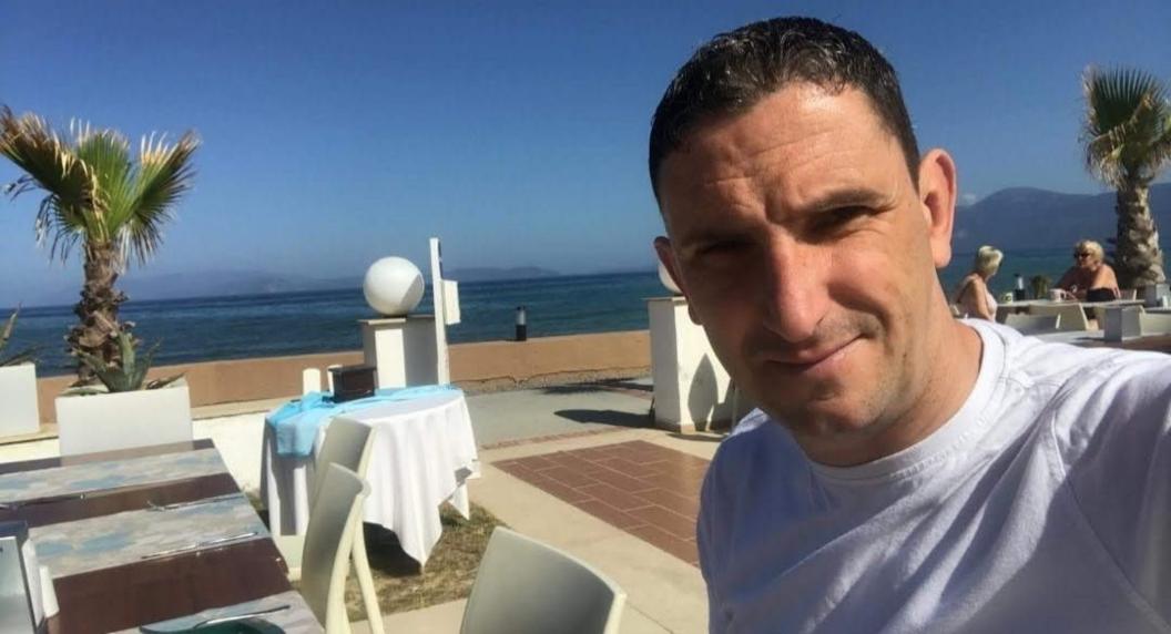 Man with dark hair in front of sea, restaurant and palm trees.