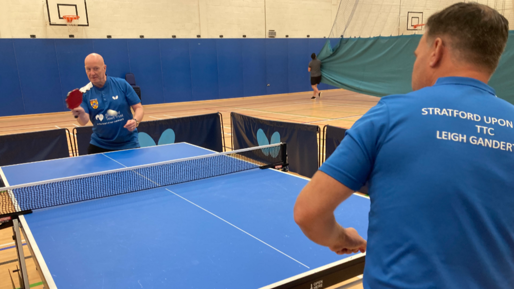 Two men playing table tennis on a blue table, with the white ball captured in mid-flight