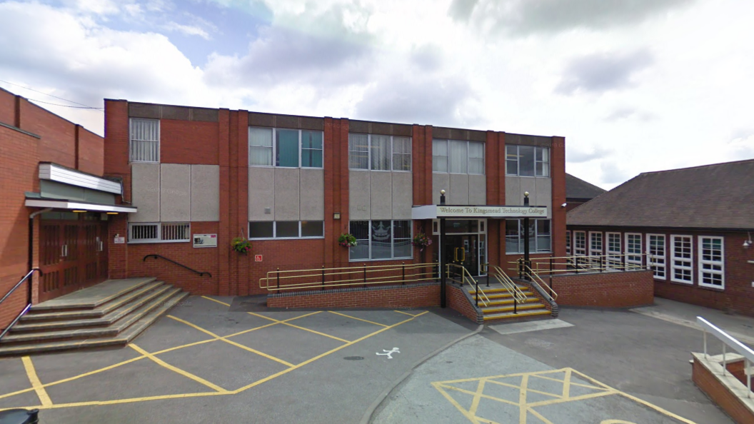 The exterior of a red-brick school building with steps and ramps leading up to the doors. There are yellow lines marked out on the car park in front of the building.