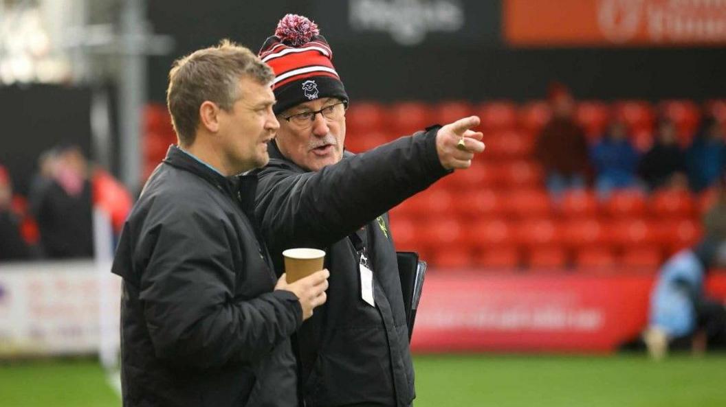 Alan Long, wearing glasses, a Lincoln City red, white and black hat and a black coat, pointing with his right hand. He is stood next to a man who is also wearing a black coat and holding a coffee cup in his right hand. They are stood on a football pitch. Red stadium chairs are blurred in the background. 