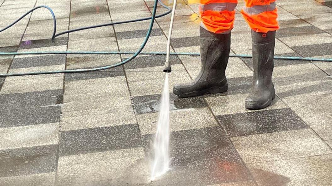 Water is being applied with pressure from a pipe to a black and white checked pavement. A par of boots and high vis orange trousers worn by the operator are also in the shot.
