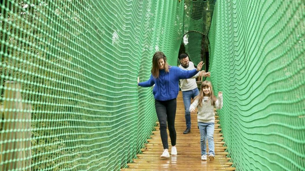 A woman, man and girl walking down a wooden walkway lined with green netting. The woman is wearing a blue jumper and black trousers and has one hand on the netting. The girl is next to her and is looking ahead. 