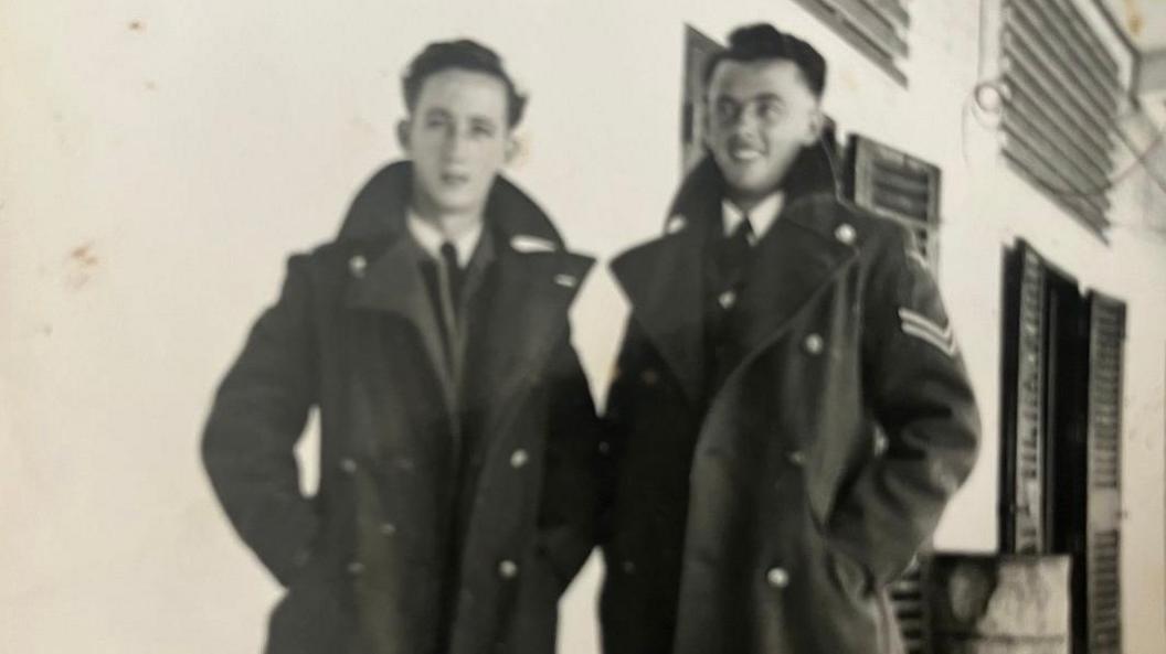 Two military servicemen stand side by side in an old photograph from the 1940. The image is black and white, both men are wearing long dark coats with military insignia on the sleeves.They are standing in front of a white wall, with a door visible to their right. It appears to have shutters.