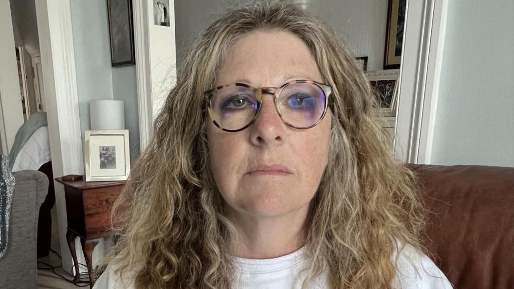 A lady with curly blond hair wearing a white top sits on a brown leather sofa in her lounge
