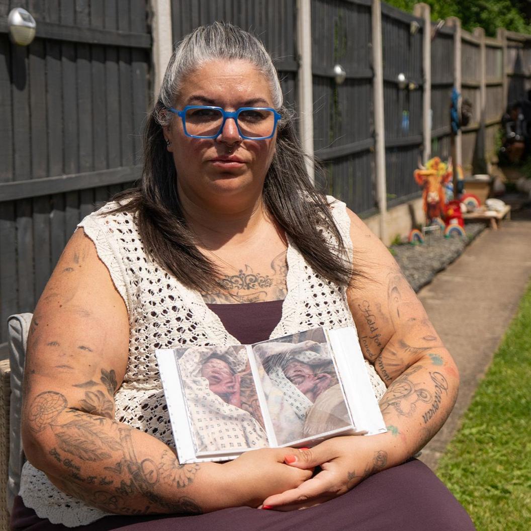 Natalie's Needham, who wears blue glasses, sits in her garden and holds two pictures of Kouper. A fence can been seen behind her.