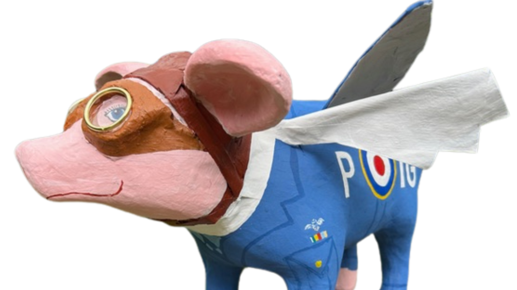 A painted pig with a wry smile, painted to have a smart blue uniform with RAF insignia. He also has a pair of old-fashioned flying goggles and a hat. Plane wings are attached and some tissue has been made to stick out from his neck, as if it is a scarf flying in the wind.