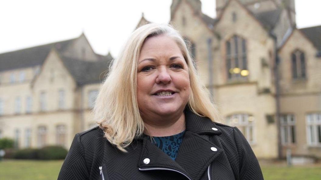 Joanna Hudson is standing in front of a large Victorian brick building, which is blurred in this image. She has shoulder-length blond hair and is wearing a blue top and a black jacket