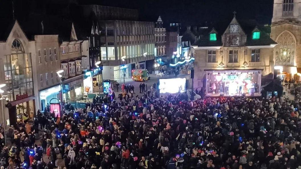 A large crowd of people in Cathedral Square, Peterborough. You can see people wrapped up in coats and hats with shops, the Guildhall and St John's Church in the background