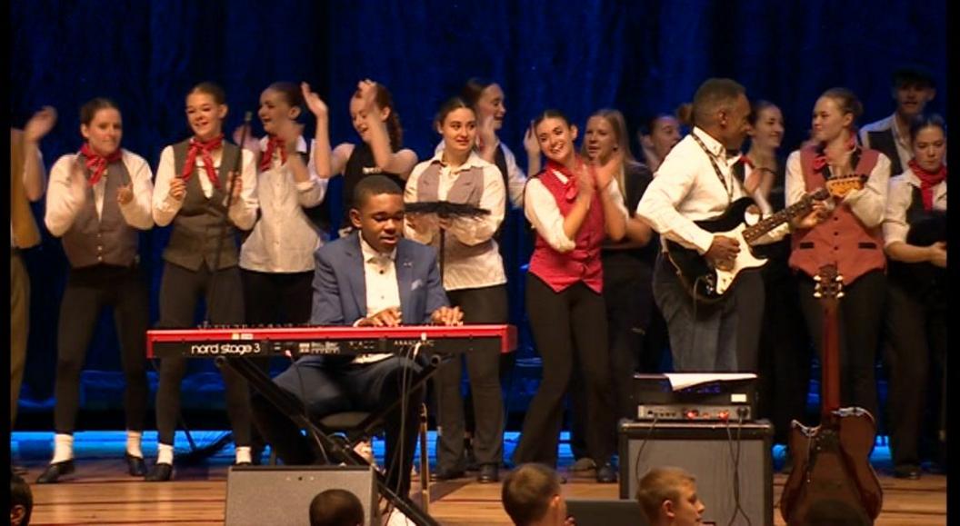 Students from Venturers Academy stage a performance at the Bristol Beacon dressed in red and grey waistcoats while a student plays keyboard. 