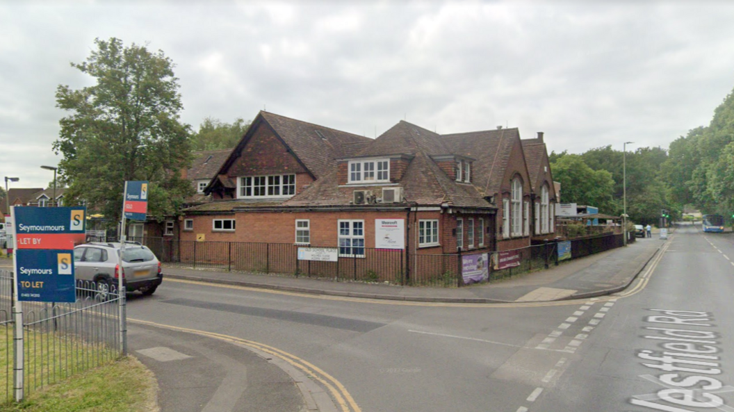 A brick community centre on the corner of a road with trees in the distance. It is an overcast day. 