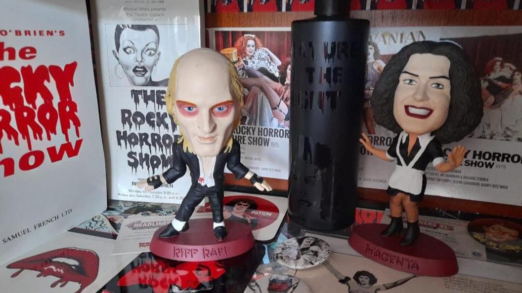 A display of 'The Rocky Horror Show' memorabilia featuring figurines of Riff Raff and Magenta, surrounded by posters, flyers (including one from King's Road Theatre), a black bottle with the phrase 'DON'T DREAM IT BE IT', and various themed buttons, cards, and images