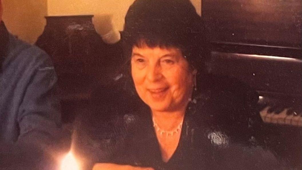 Rita Grimshaw is smiling while sitting with others at a table and looking at a lit candle. She has short black hair and is wearing a smart black top and silver necklace. There is a piano behind her in the room with white walls, that appear yellow in the candlelight. The photo is grainy and looks to be a photo of a physical picture.