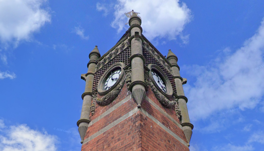 A picture angled up at the King Edward VII Memorial Clock Tower focused on the top part which has two clockfaces visible with ornate stone carvings surrounding them.