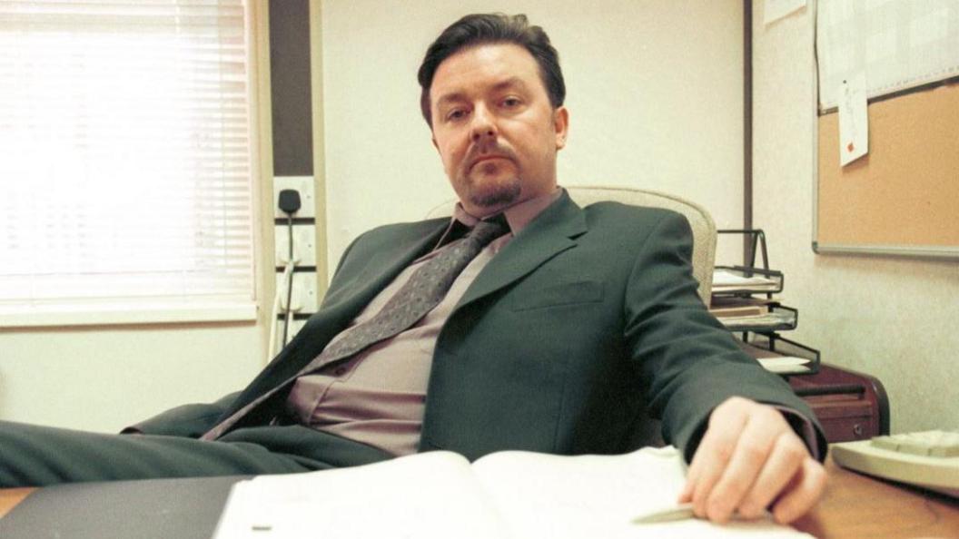 Ricky Gervais as David Brent in The Office, based in a paper merchants in Slough. He is wearing a suit and ties and slouching on a chair at a desk.