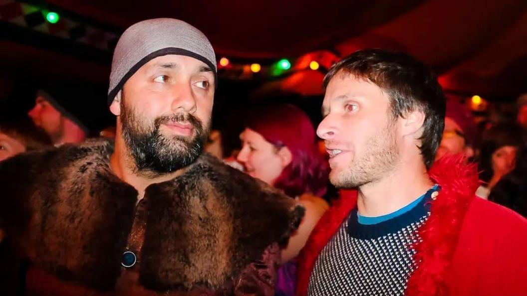 Kaptin Barrett (left) pictured with Stu (right) as they both wear fancy dress. Kaptin has a dark brown bushy beard, brown eyes and wears a hat and fur shawl. Stu has brown hair, a stubble beard and looks away from the camera. He wears a red jacket with a hair trim.