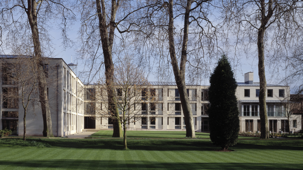 long, grey college building with elderly trees in the foreground and a well tended lawn