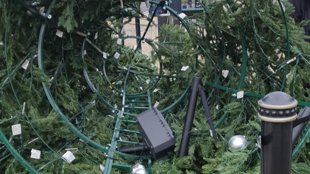 The inside of a cone-shaped artificial Christmas tree, lying on its side on a pavement. It looks like a metal-framed structure, with a white sandbag under the front section. Silver baubles are attached to the green branches. It has a lightweight webbing cordon around it and there is a black and gold bollard on the right