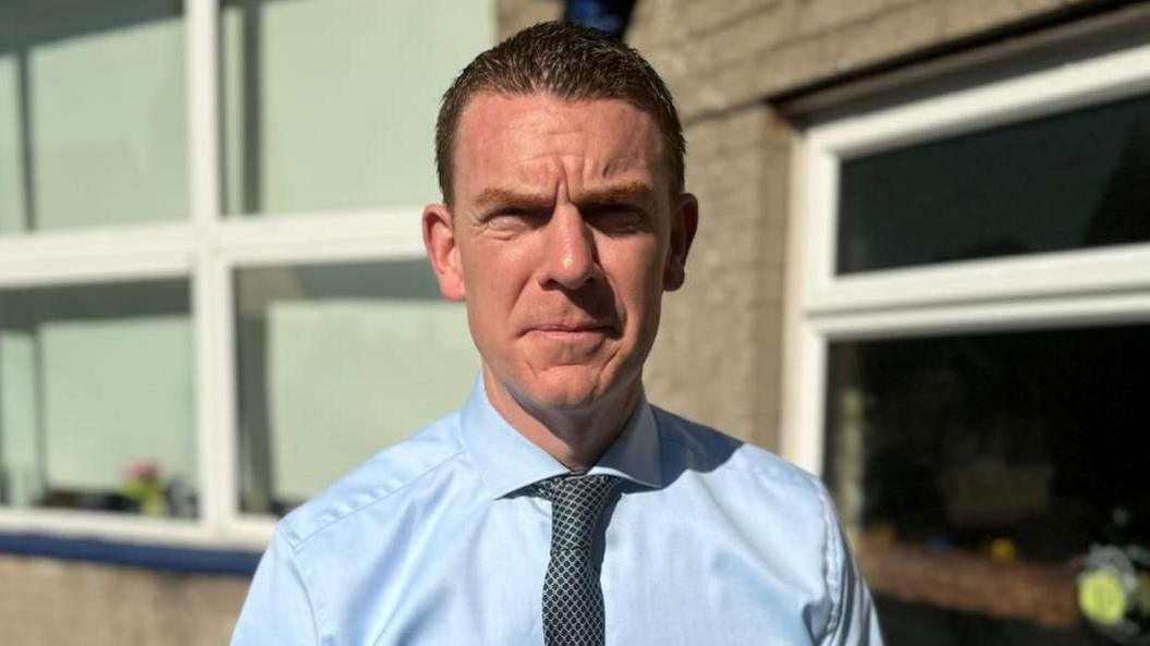 A man is looking directly into the camera. He is wearing a light blue shirt and a dark patterned tie. The background is out-of-focus, but you can make out the wall of a school with a run of windows and flowerbeds. 