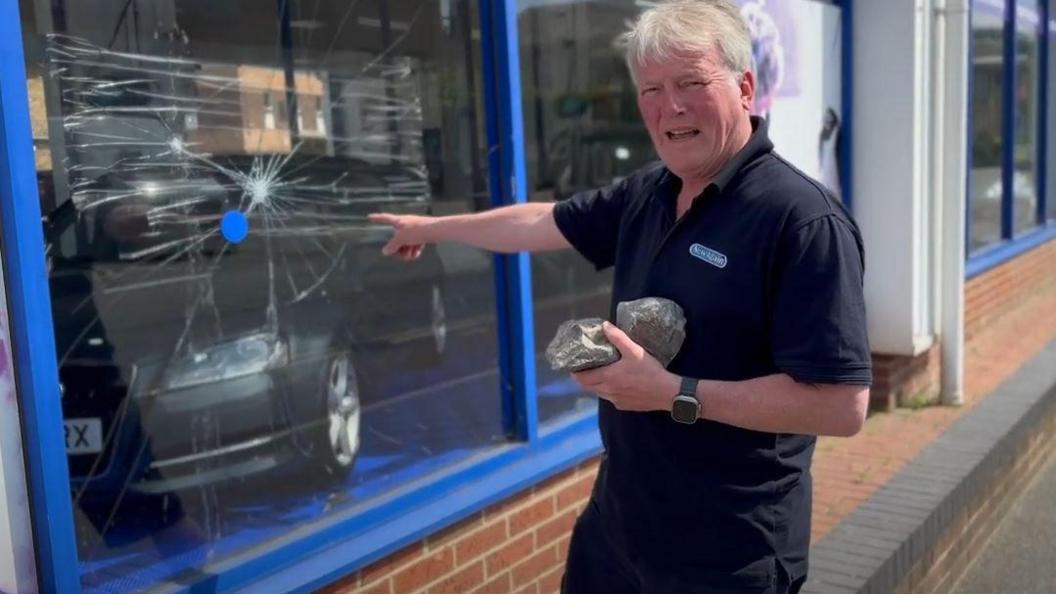 A man standing in front of a shop window with a very large crack in it He is holding two fist-sized stones