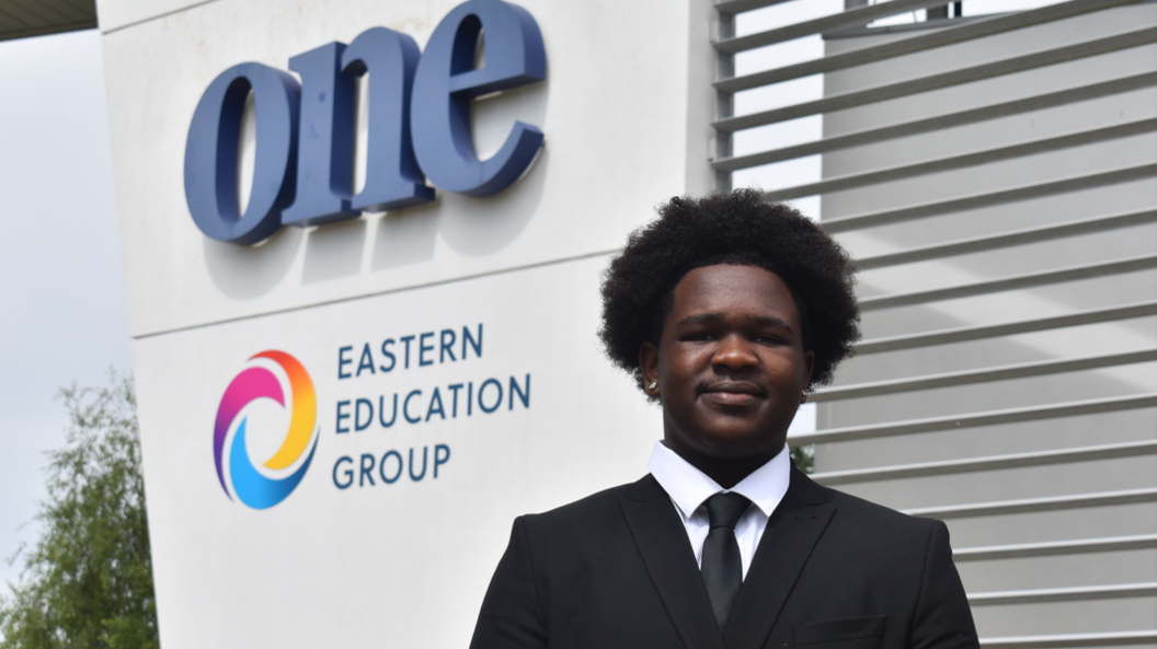DJ Mudziviri wearing a black suit jacket and tie over a white shirt. He is standing in front of Suffolk One Sixth Form College, the logo for which can be seen to the left of the image. He is looking and smiling directly into the camera