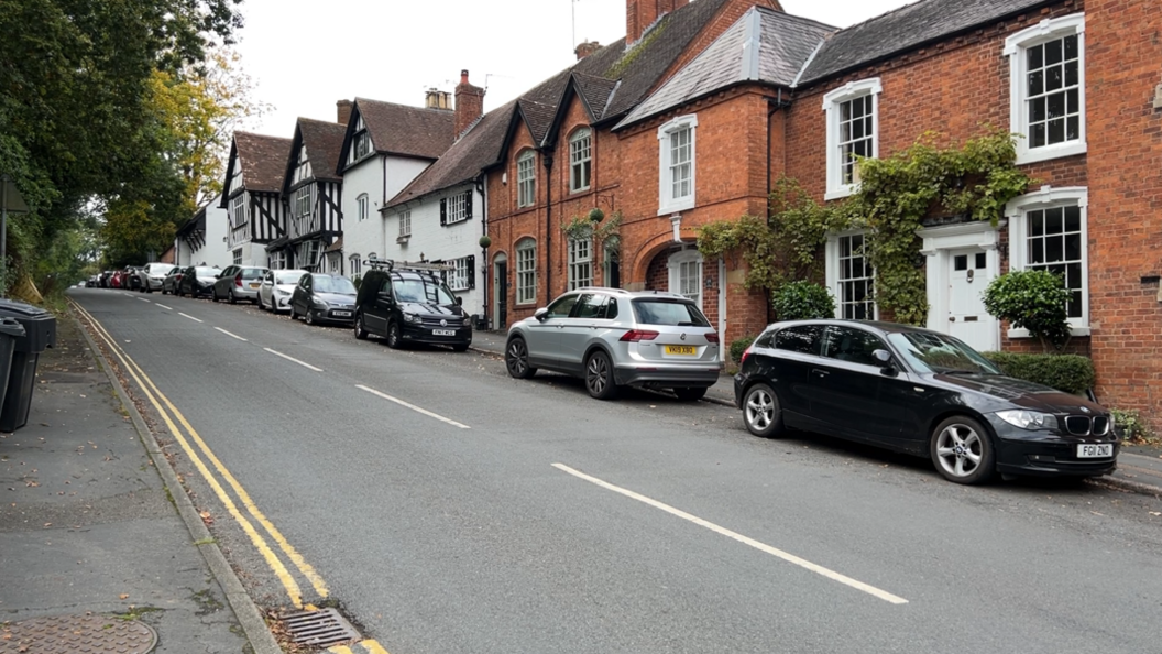 A street in Alvechurch, shows trees and a wheelie bin on a pavement on the left with cars parked and a line of older properties on the right