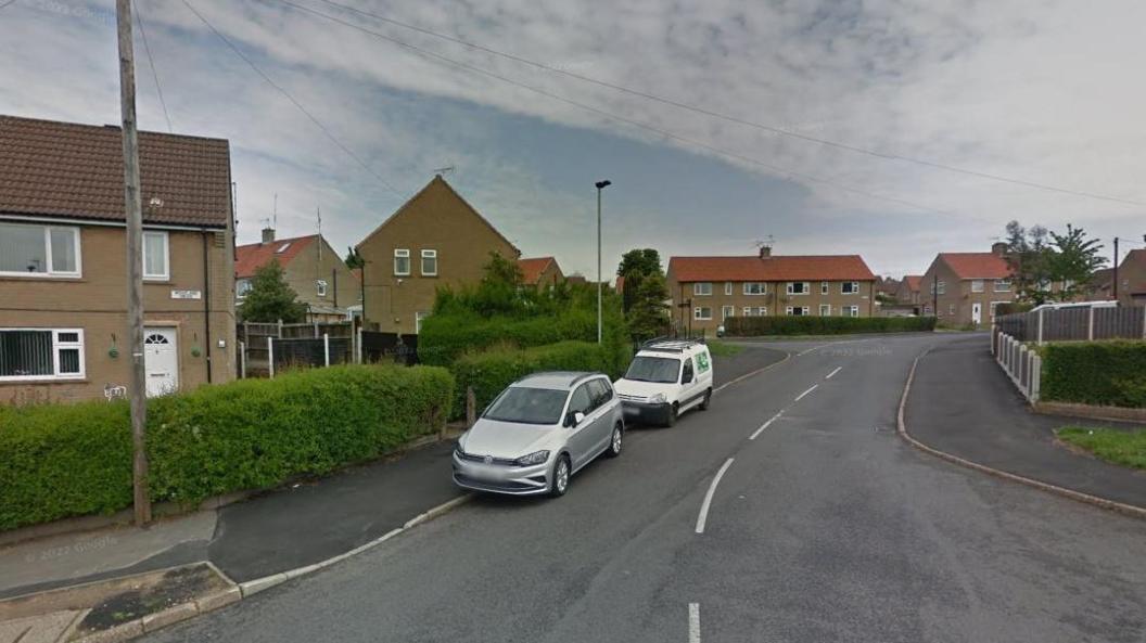 An asphalt road with white line markings leads to a series of council house. Two cars, one white, the other silver are parked straddling the road and pavement.