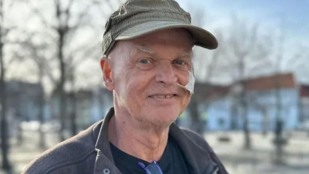 Kian Reme smiles at the camera. He is wearing a green hat and has a tube coming out of his nose. He is wearing a navy jacket and is standing in front of some buildings and trees