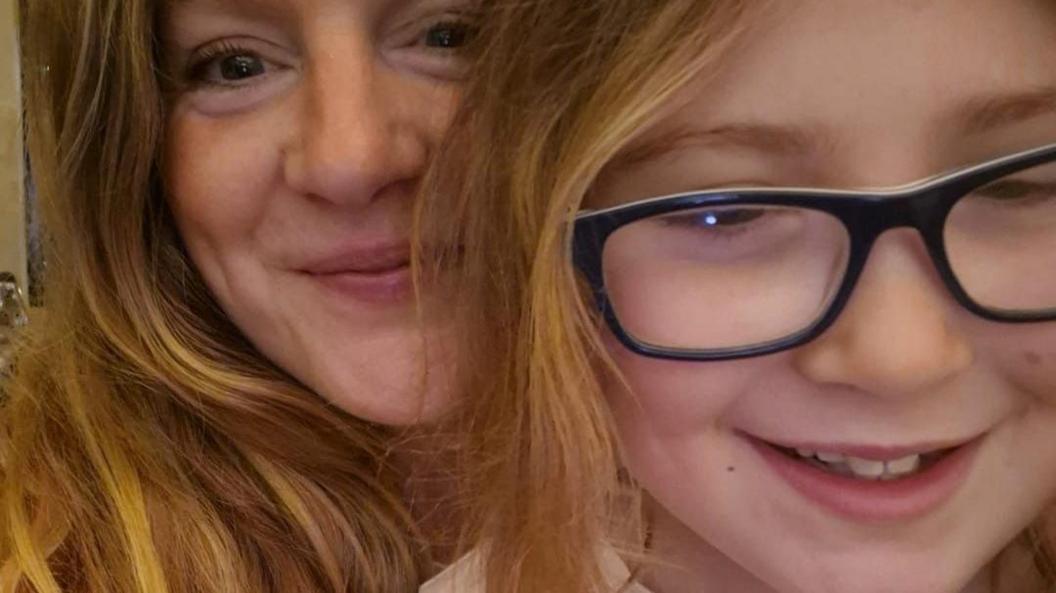 Caroline, a woman with long sandy blonde hair looking into the camera lens, smiling. Picture also features her daughter Lotus, aged nine, who also has sandy blonde hair, and is wearing navy blue spectacles.