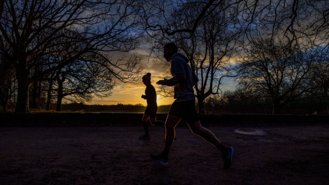 Silhoutted joggers run through a park. The sun is low in the sky, and there are trees surrounding the runners