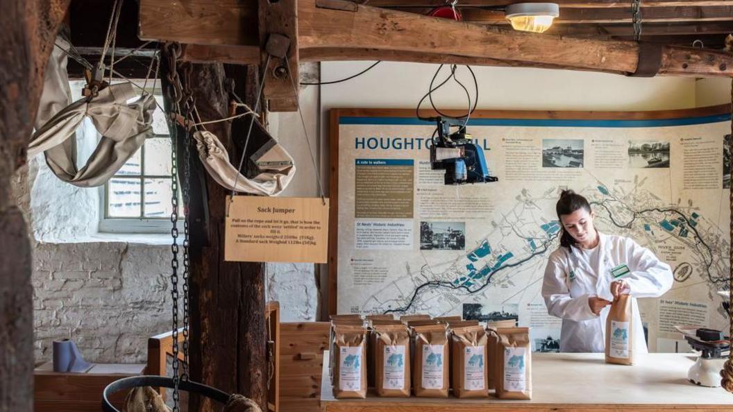 A woman in a white coat puts flour into brown paper bags inside Houghton Mill.